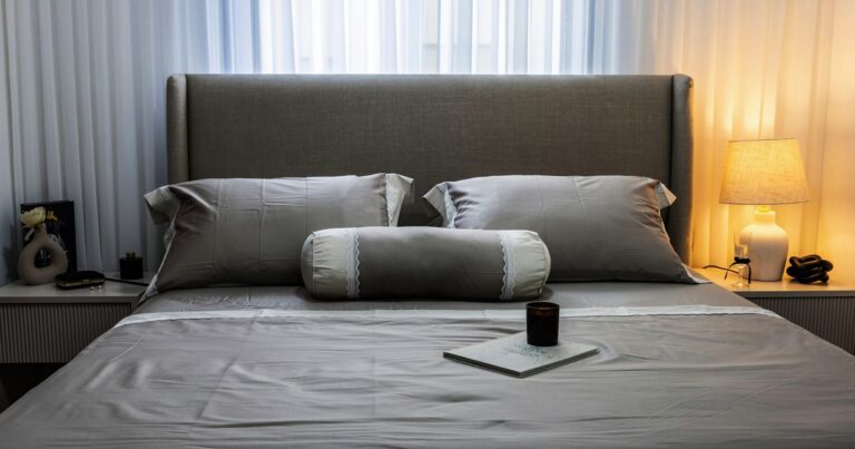A made bed with a candle on the covers and a lamp lit beside it