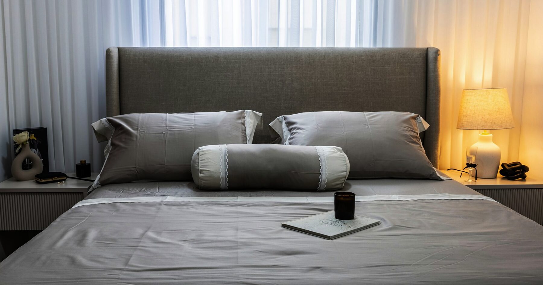 A made bed with a candle on the covers and a lamp lit beside it