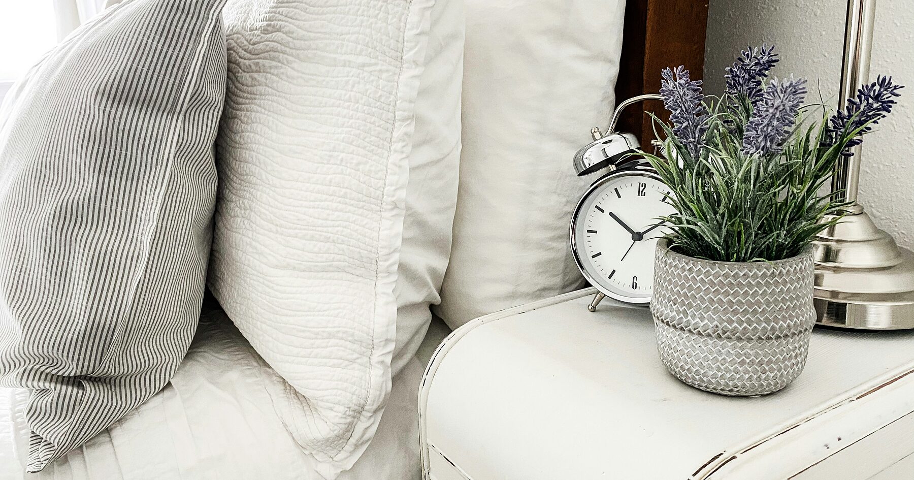 Pillows on a bed beside an alarm clock and a pot of lavender