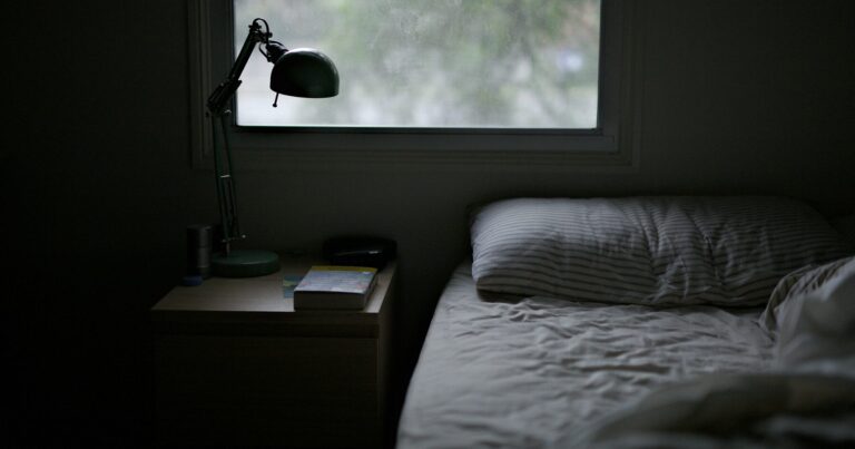 A dark, cool bedroom with a small window and an unlit bedside lamp