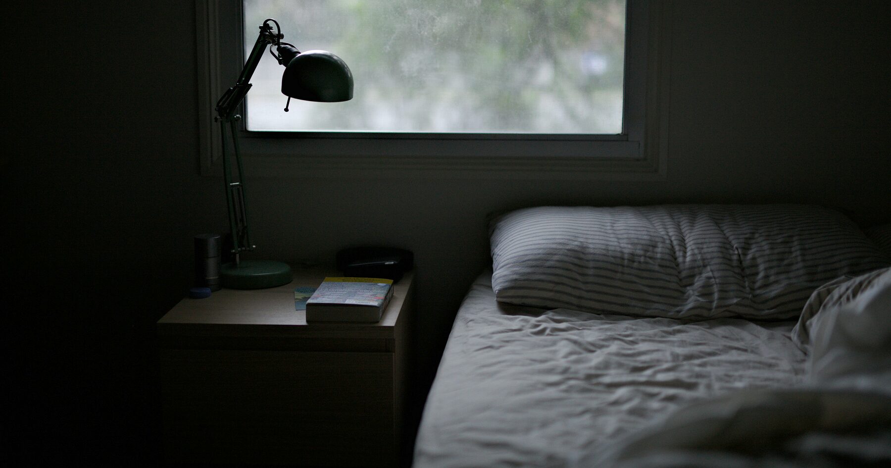 A dark, cool bedroom with a small window and an unlit bedside lamp